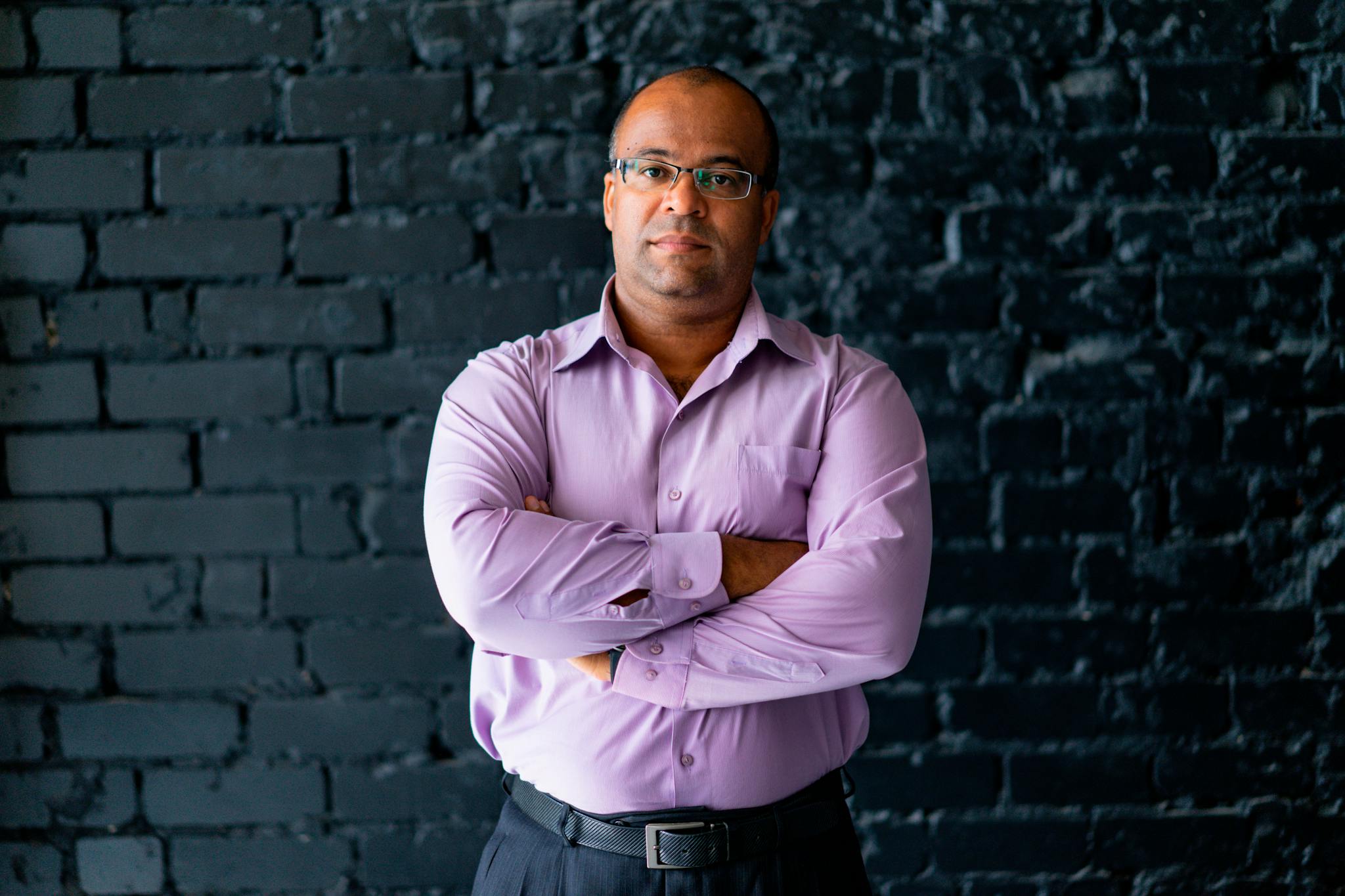 Portrait of a confident man in eyeglasses standing with arms crossed against a brick wall.